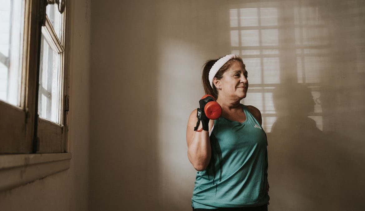 Middle-aged woman in workout attire lifting a dumbbell indoors, promoting fitness and healthy lifestyle.